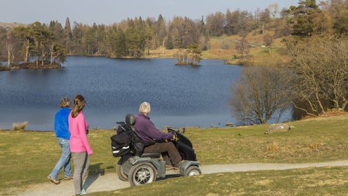 A group of people, one in a mobile tramper, explore along the side of a lake. The lake is surrounded by rough grass and mixed woodland.